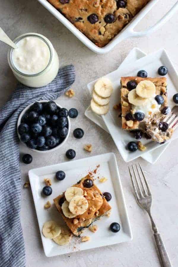 aerial view of Blueberry Banana Walnut Muffin Bars on white plates with a bowl of blueberries and a jar of yogurt