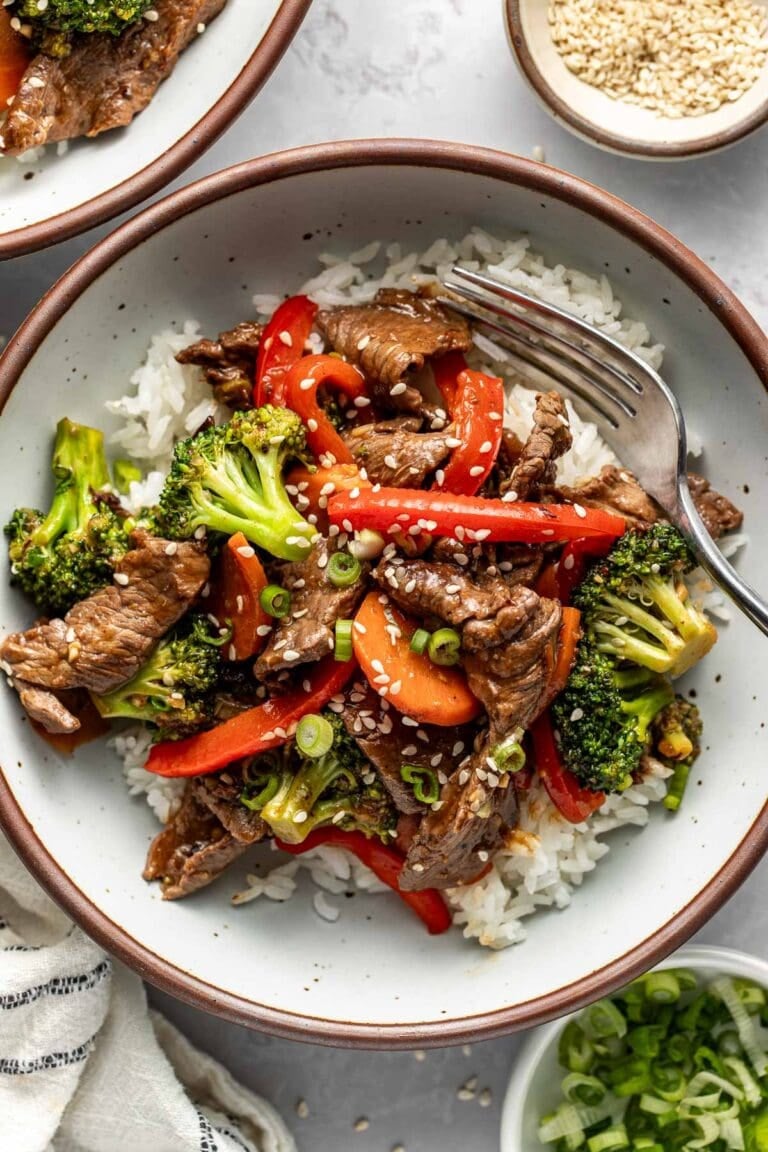 Overhead view of a serving of steak stir fry with red bell peppers, broccoli, and carrots over top of white rice and garnished with sesame seeds.
