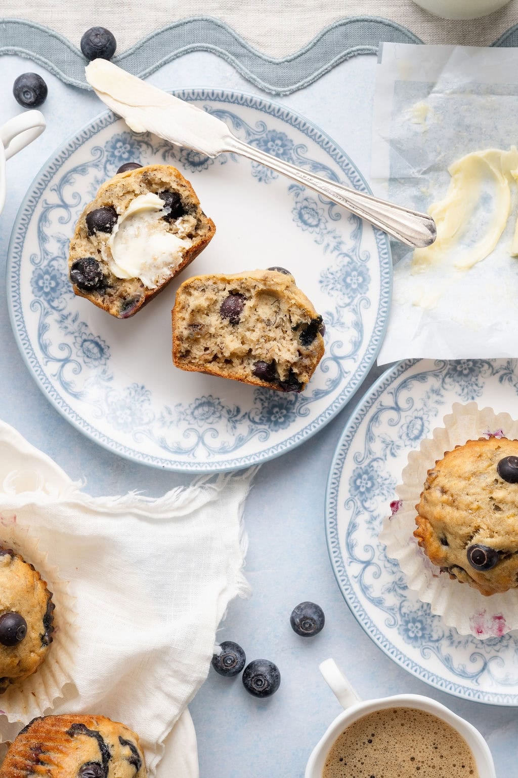 Overhead view of a Banana Blueberry Muffin cut in half on a plate and smeared with butter. 