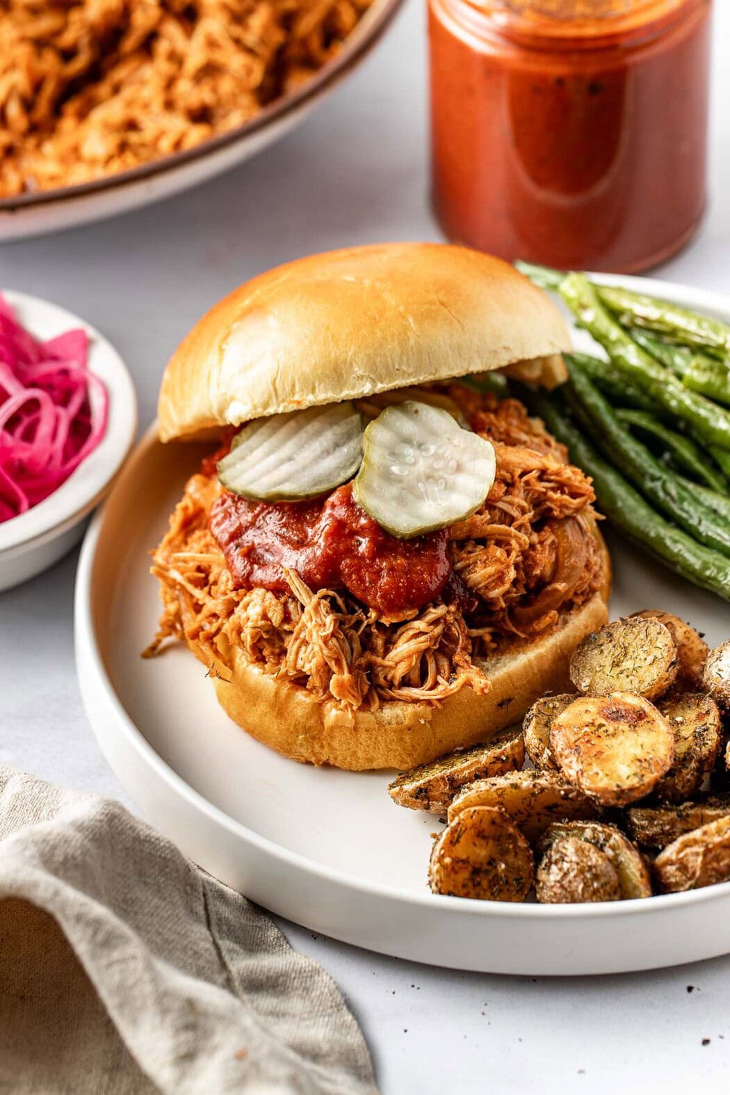 Close up view of a plate with a BBQ Pulled Chicken sandwich on it next to roasted potatoes.