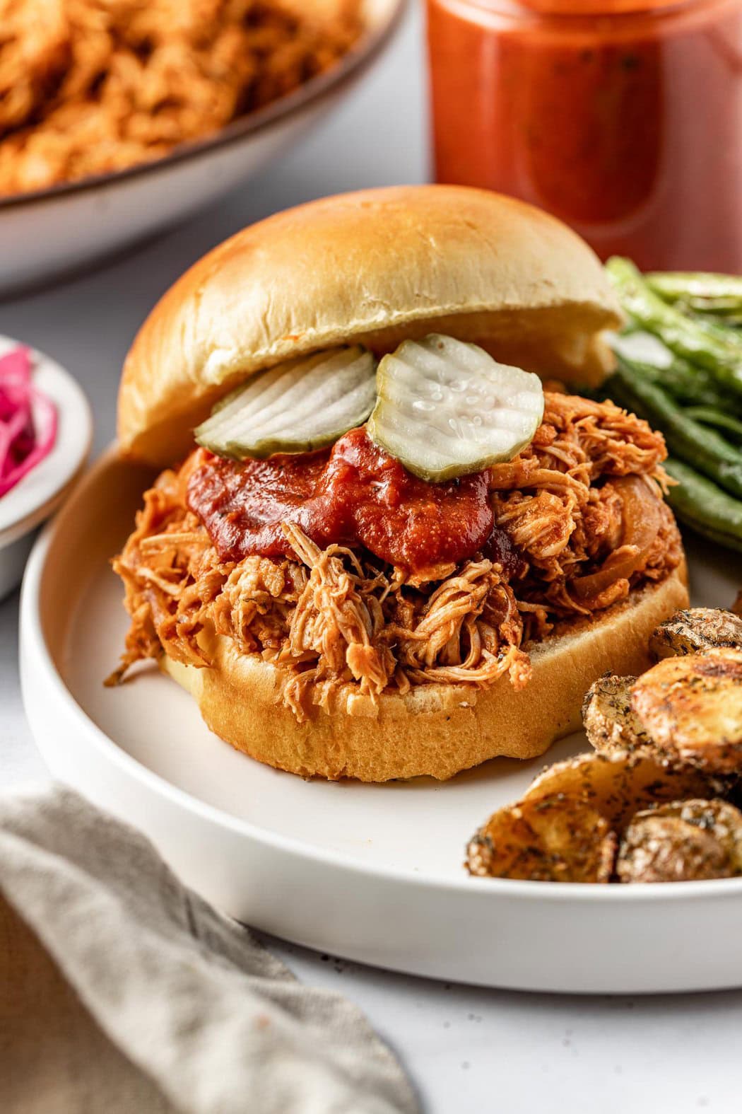 Close up view of a plate with a BBQ Pulled Chicken sandwich topped with sliced pickles and BBQ sauce. 