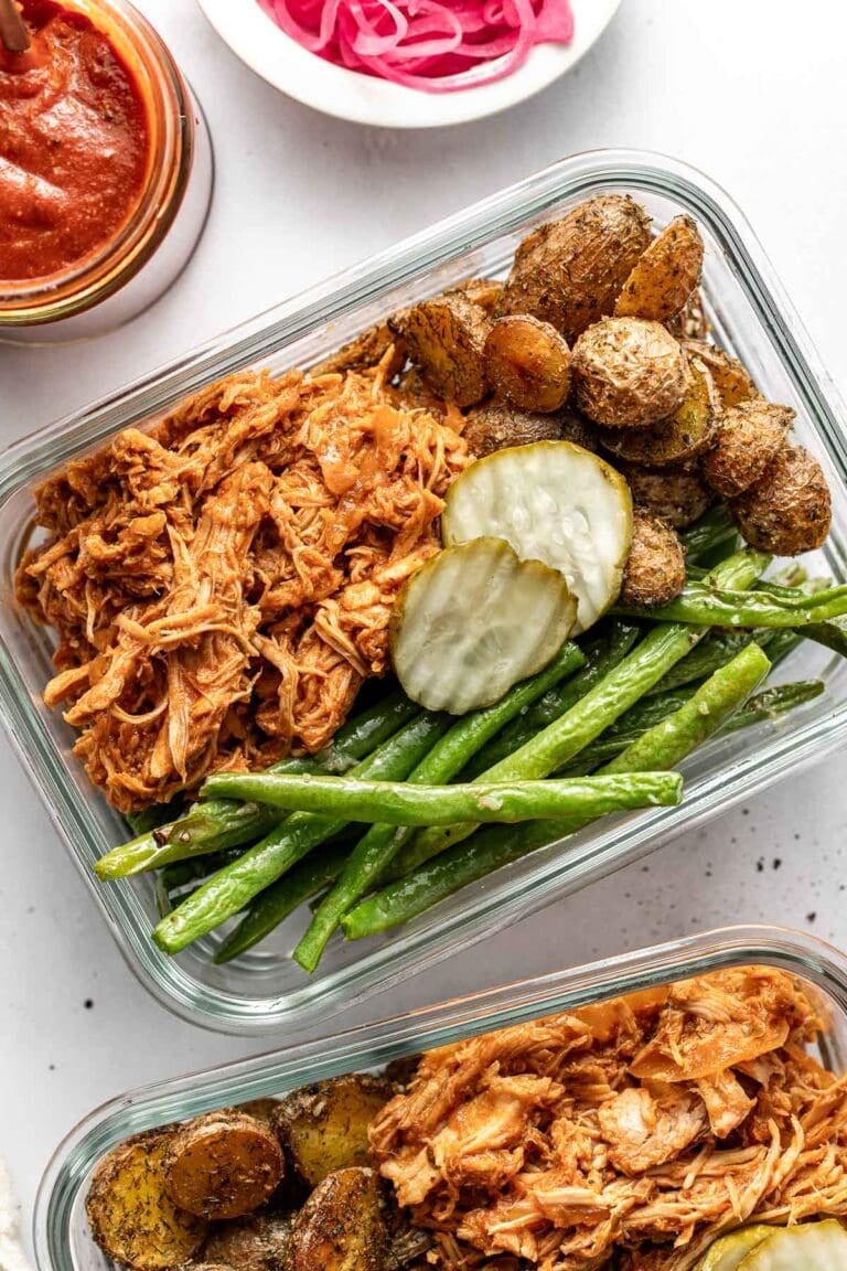 Overhead view of two glass meal prep containers filled with BBQ pulled chicken, roasted potatoes, and green beans topped with pickle slices.