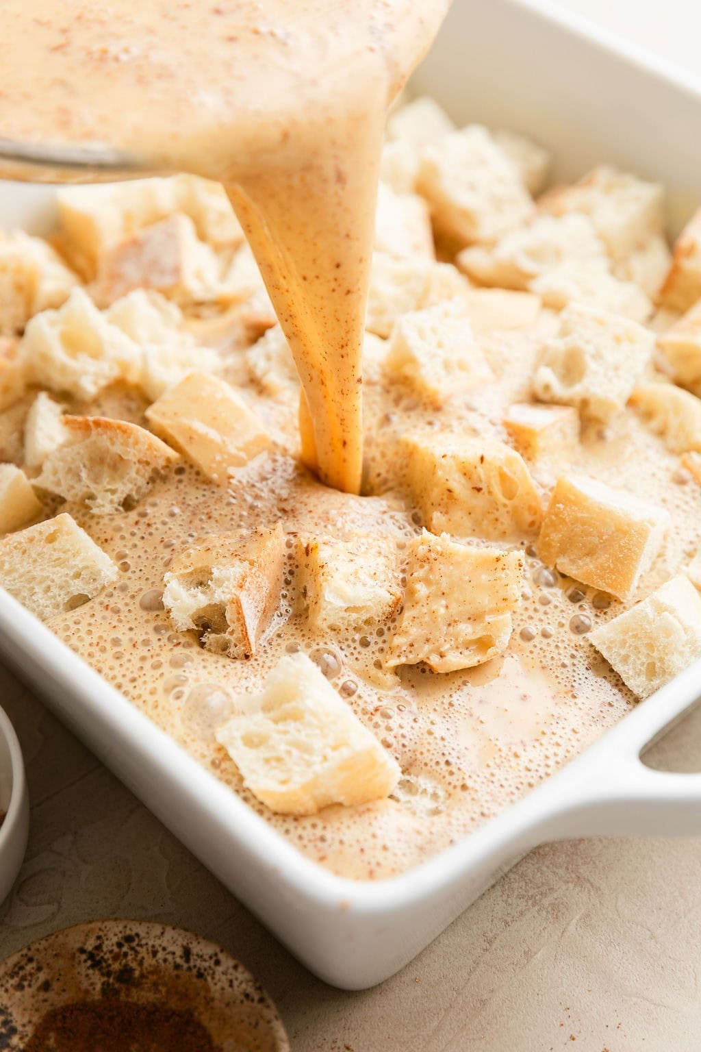 Close up view of a baking dish of bread pieces with an egg mixture being poured over the top.