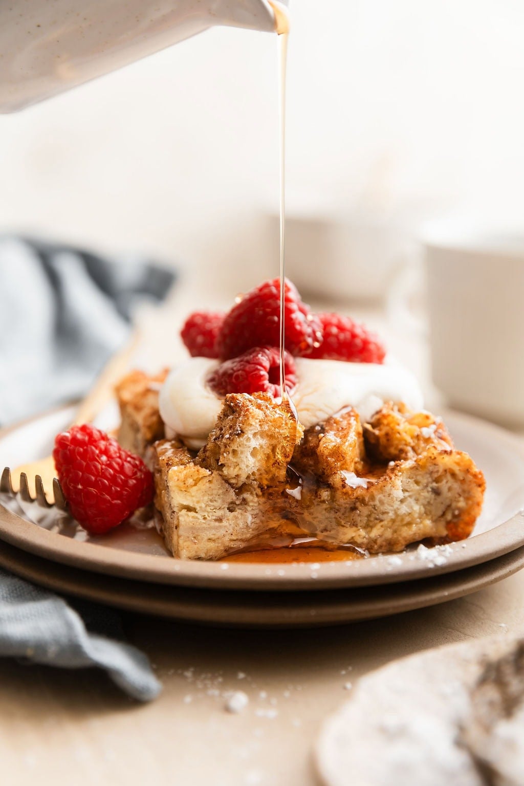 Close up view of a piece of French Toast Casserole being drizzled with maple syrup and topped with whipped topping and fresh raspberries.