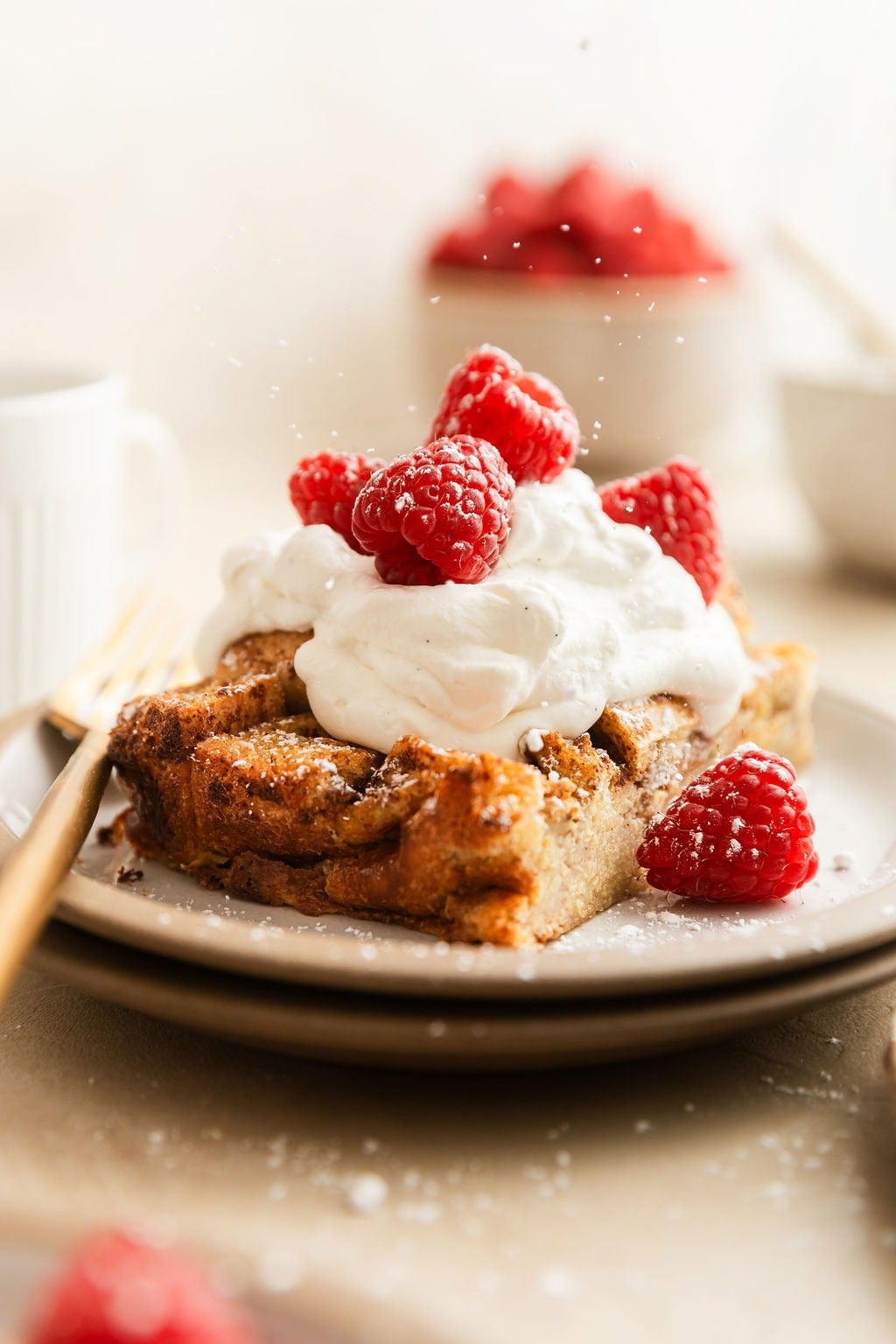 Close up view of a plate filled with a piece of French toast casserole topped with whipped topping, fresh raspberries and sprinkled with powdered sugar.