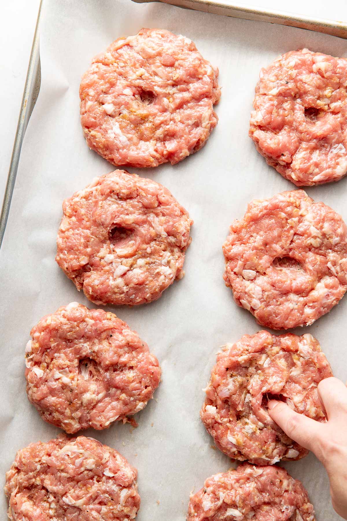 Overhead view of a parchment lined sheet pan with turkey patties on it showing a hand pushing an indentation in the middle. 