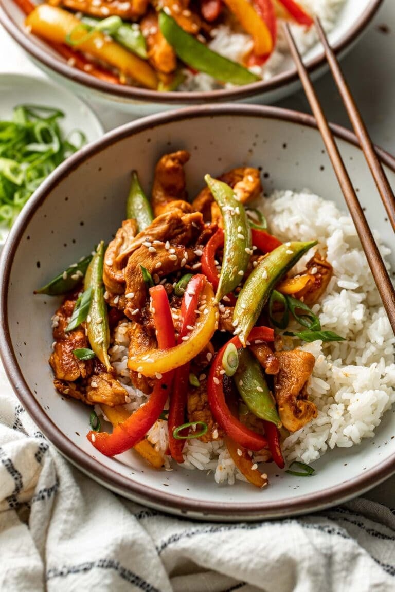 Close up view of a bowl of chicken stir fry on top of a bed of rice topped with sesame seeds. 