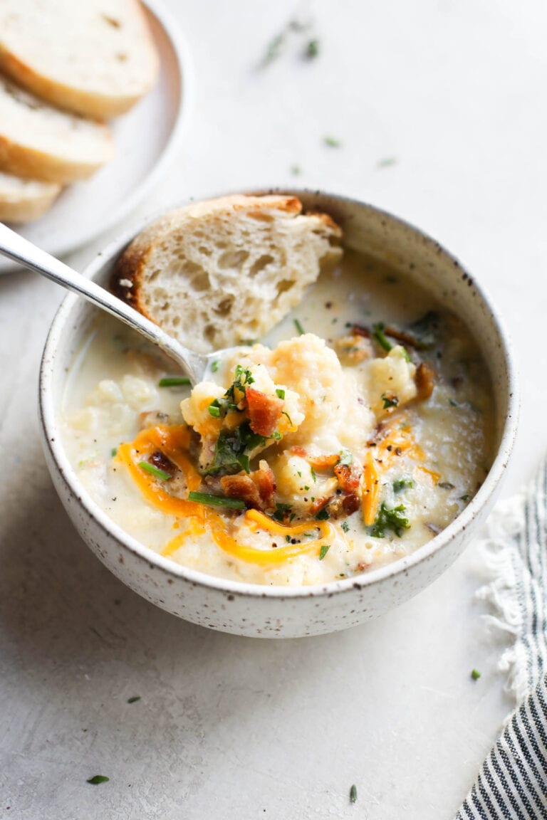 A white bowl of cauliflower soup topped with melted cheese and a piece of bread on the side. 