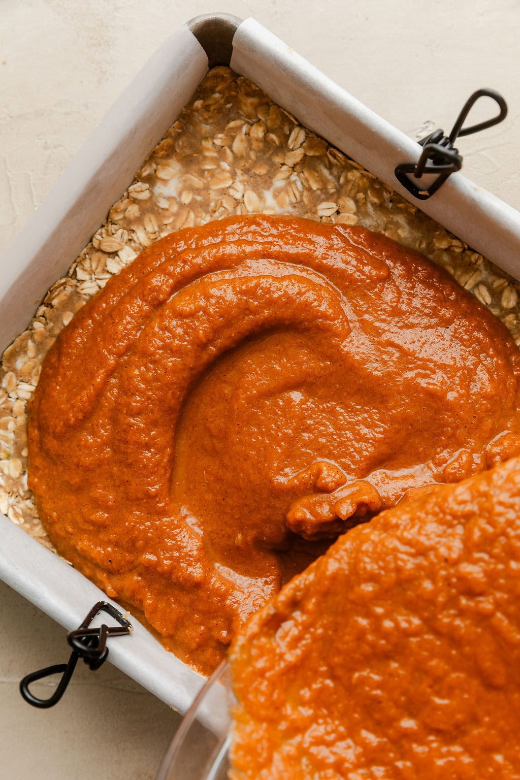 Overhead view of a pumpkin mixture being poured on top of a oat crumble in a parchment lined baking dish. 