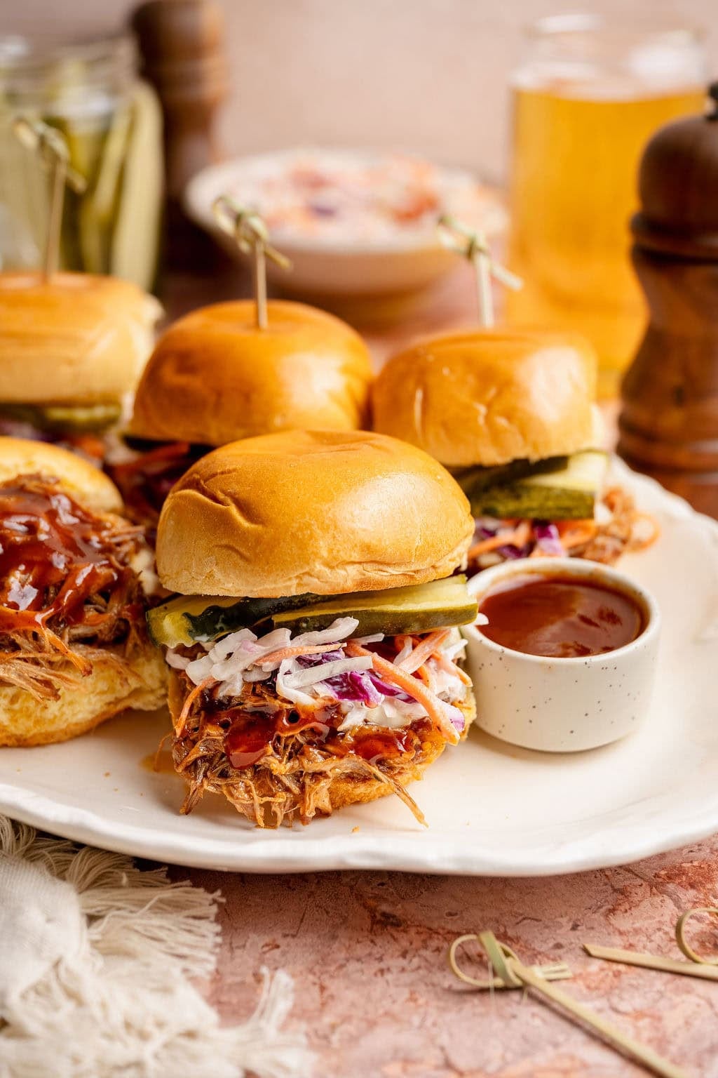 Close up view of several Pulled Pork Sliders topped with coleslaw and pickle slices with a side of BBQ sauce
