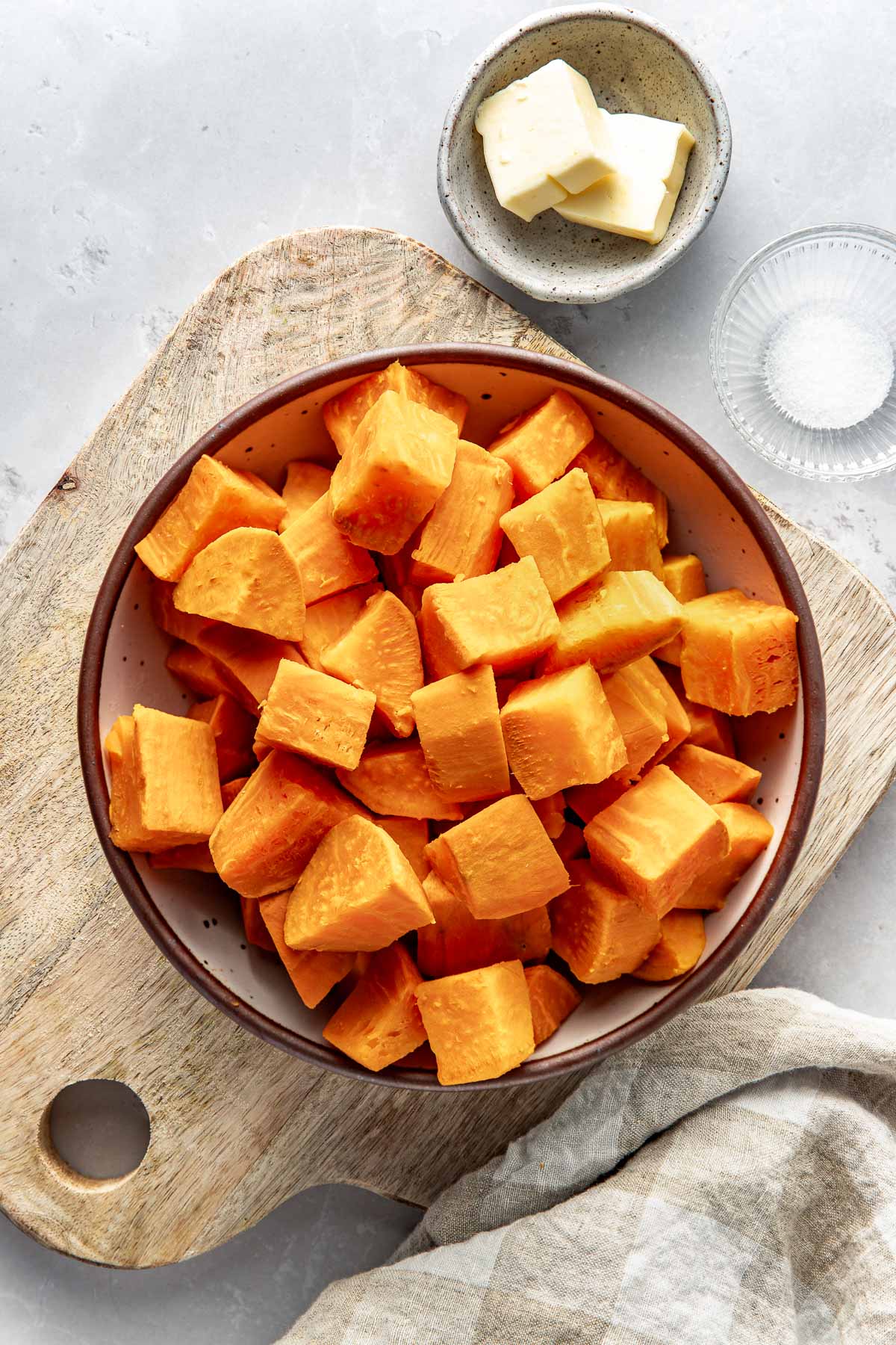 Overhead view of a bowl of diced sweet potatoes on a wooden cutting board with butter on the side in a small dish. 
