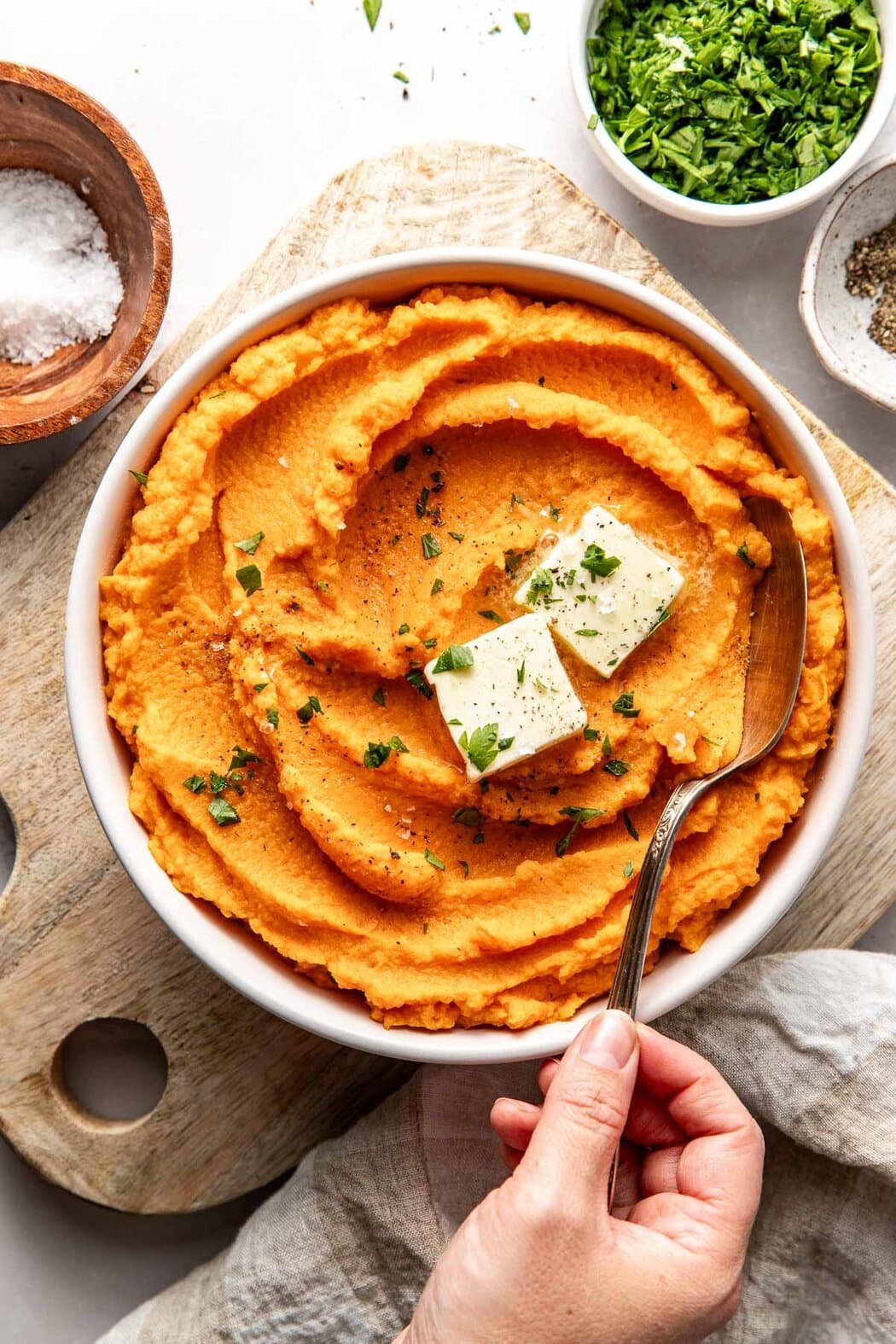 Overhead view of a bowl of Mashed Sweet Potatoes with a hand using a metal spoon to scoop a spoonful. 