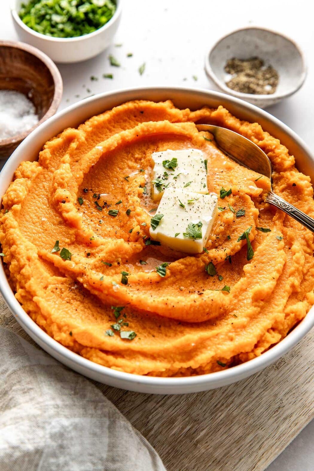Close up view of a bowl filled with vibrant orange Mashed Sweet Potatoes topped with butter and fresh herbs. 