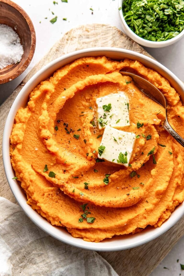 Overhead view of a bowl of vibrant orange Mashed Sweet Potatoes topped with butter and fresh herbs. 
