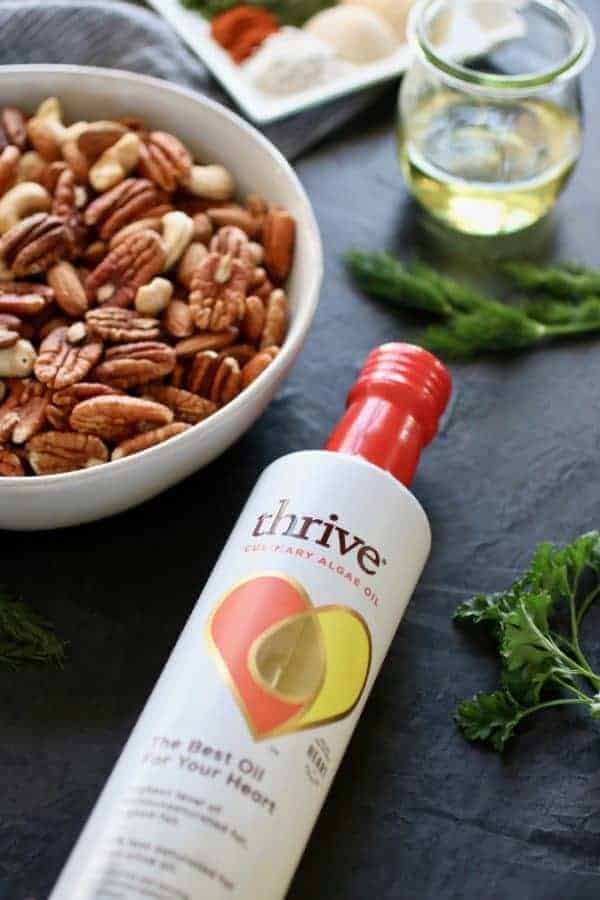 Ingredients for Ranch Roasted Mixed Nuts including Thrive Culinary Algae Oil, a bowl of raw nuts, fresh herbs, clear jar of oil and small plate of ranch spices. Photo taken on a dark background.