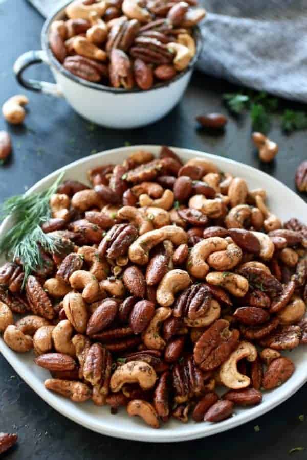 Ranch Roasted Mixed Nuts on a white plate with dill and a cup of mixed nuts in the background. Photo taken on a dark background.