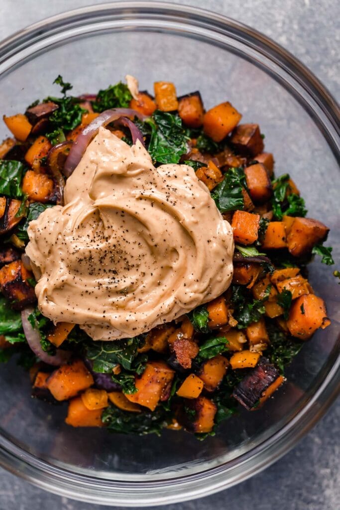 An overhead view of sautéed sweet potatoes, kale, red onion, and bacon in a bowl with chipotle lime mayo resting on top.