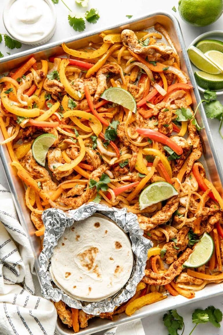 Overhead view of a sheet pan filled with fajita seasoned chicken and peppers with tortillas on the side. 