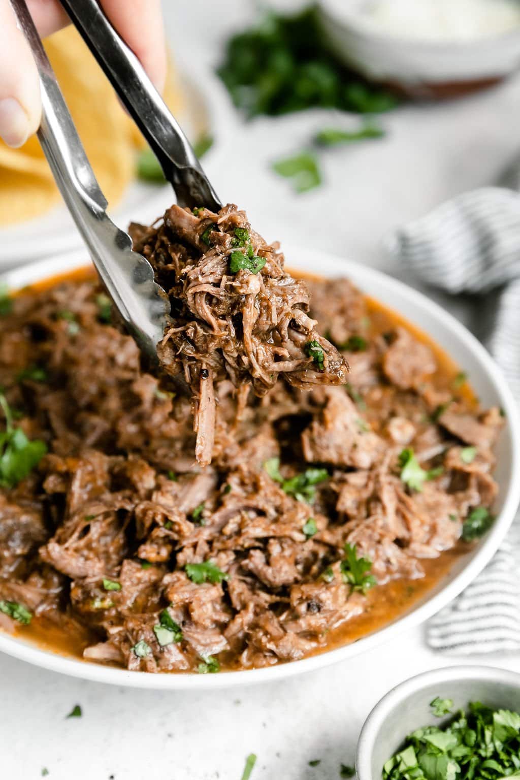 Tongs holding up serving of slow cooker beef barbacoa from white bowl of barbacoa.