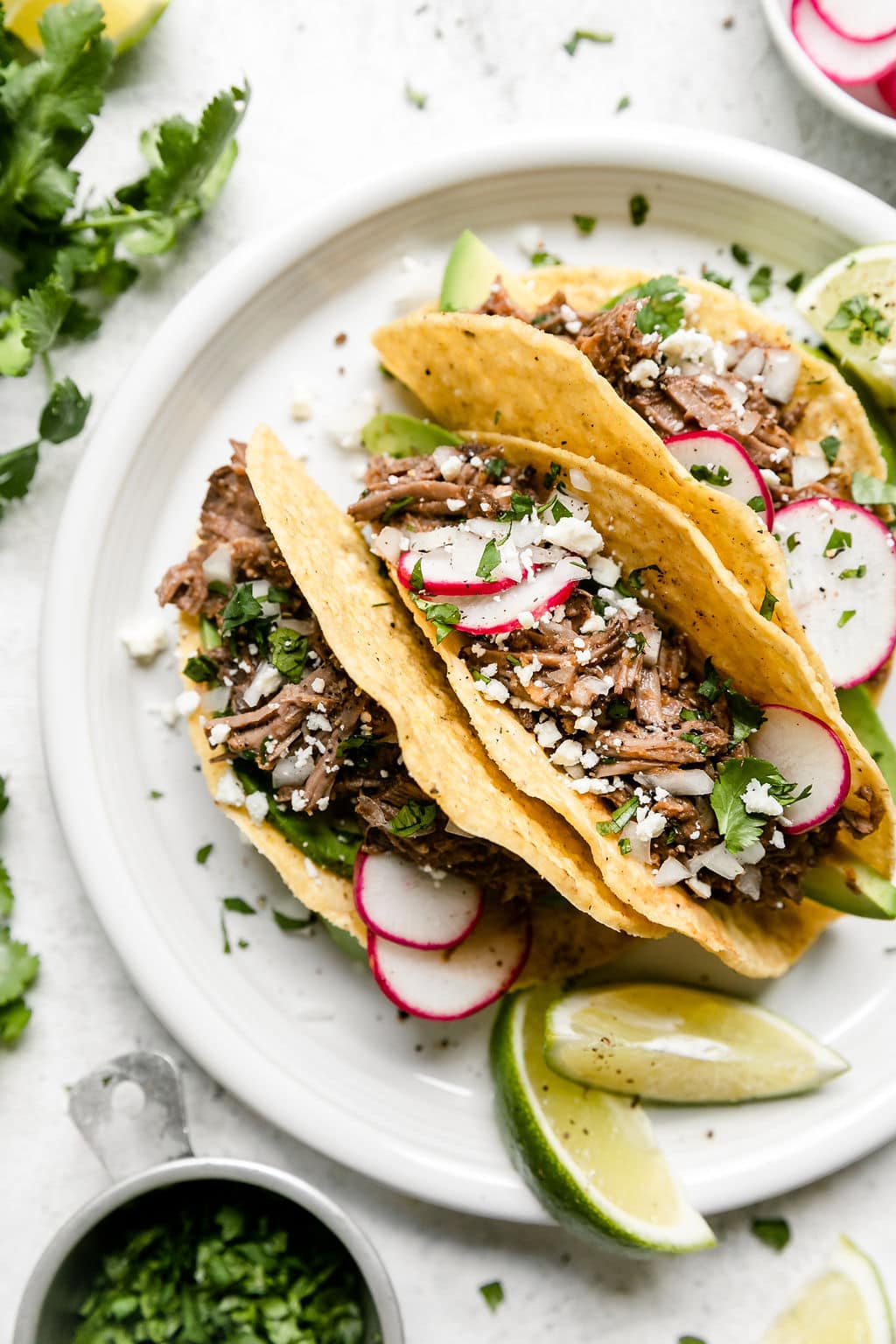 Overhead view white plate with three hard shell tacos stuffed with slow cooker beef barbacoa and sliced radishes.