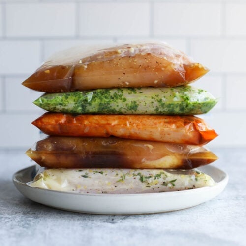 Five different chicken marinades in ziplock bags with chicken breasts stacked up on each other on a white plate.