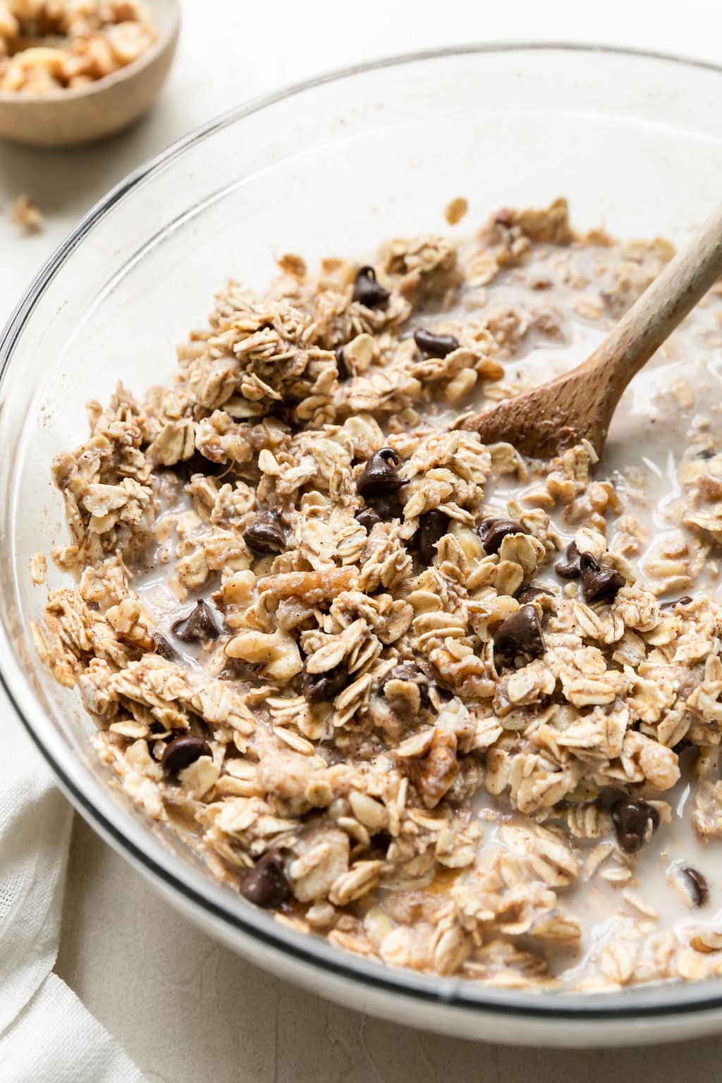 Banana chocolate chip baked oatmeal batter being stirred with a wooden spoon in clear glass mixing bowl.