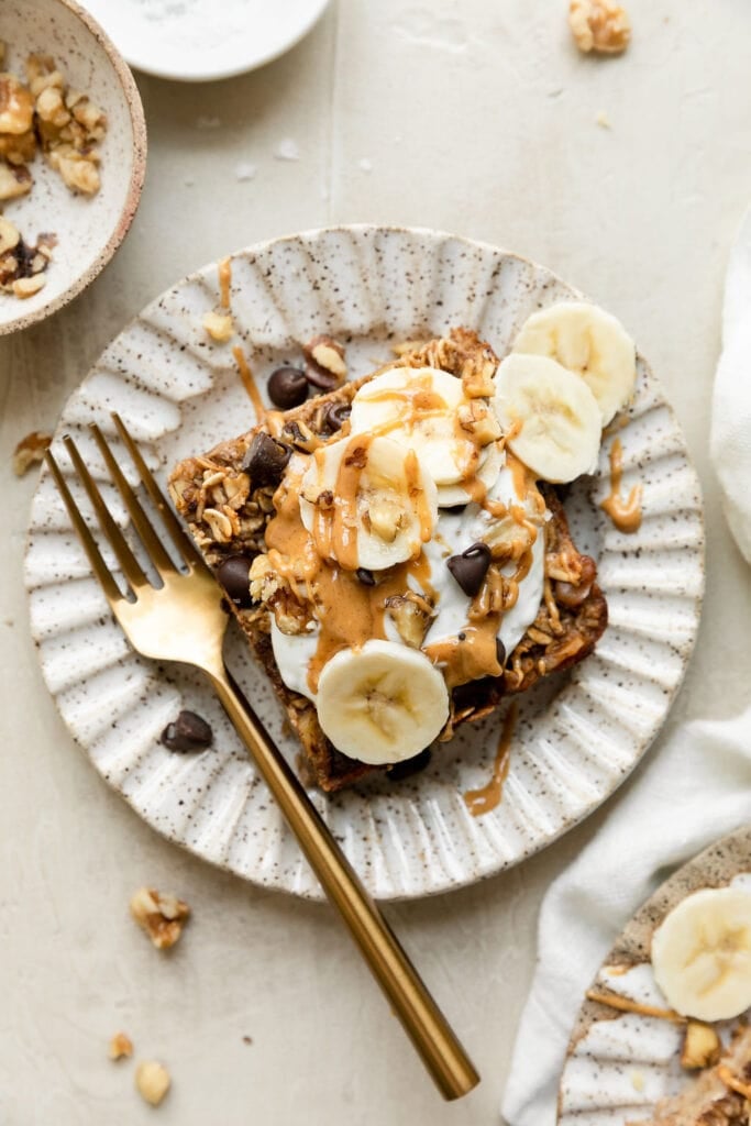 Overhead view square serving of banana chocolate chip baked oatmeal topped with yogurt, peanut butter and banana slices. 