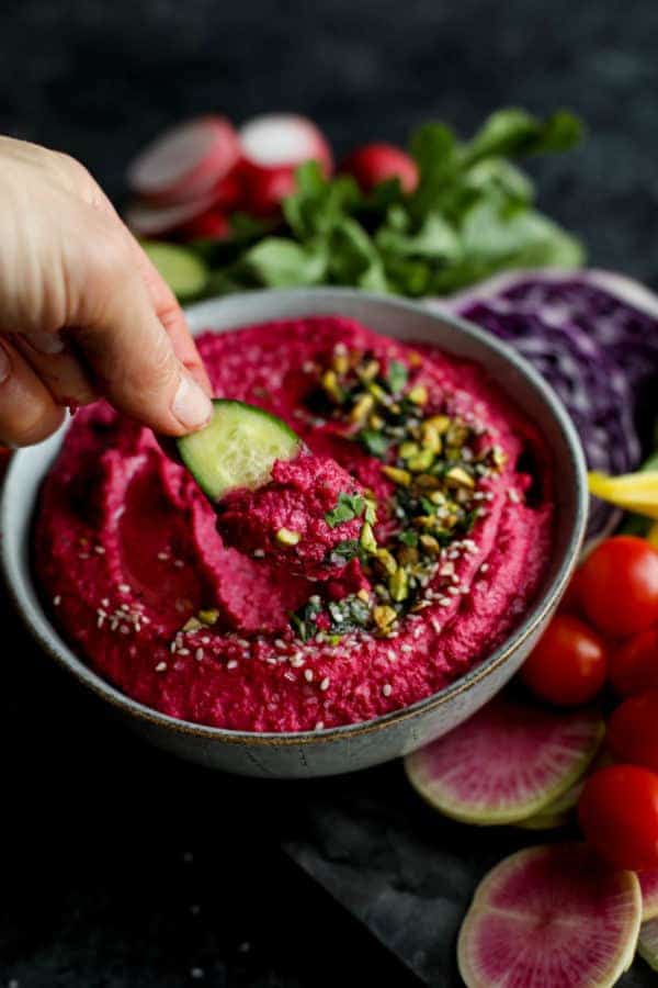 A hand dipping a cucumber slice into a bowl of Beet Hummus