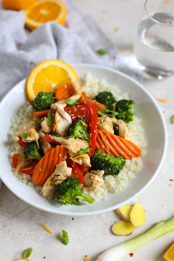White plate with stir fry chicken with carrots, broccoli, cauliflower rice and an orange garnish with a glass of water. 1 of 10 Quick and Easy Dinner Ideas.