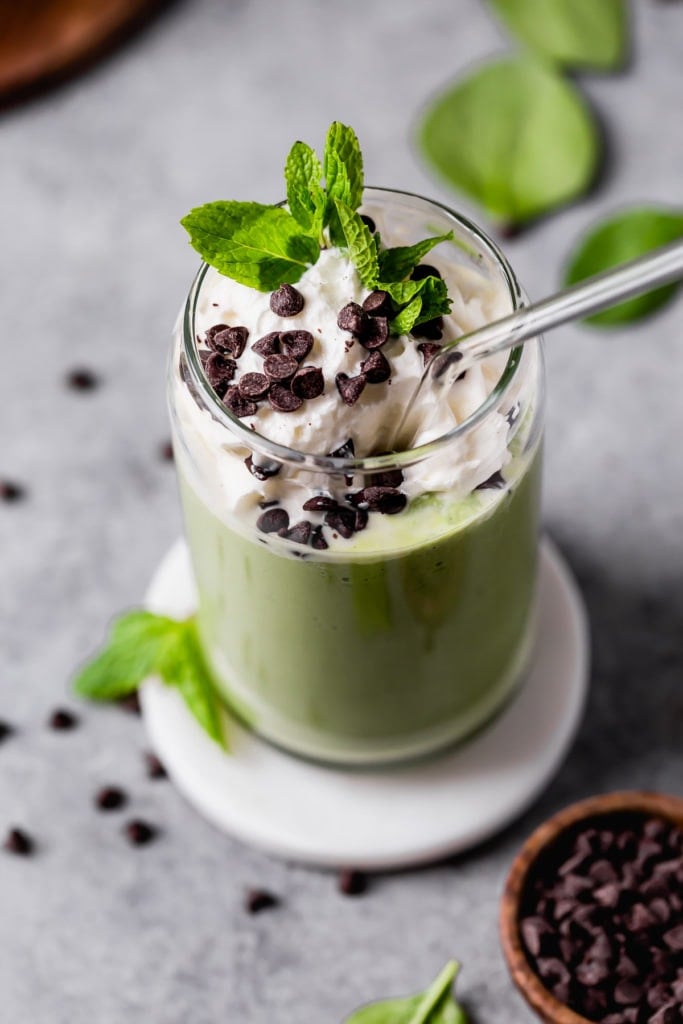 A green mint and chocolate chip milkshake in a tall glass topped with whipped cream, fresh mint, and sprinkled with mini chocolate chips