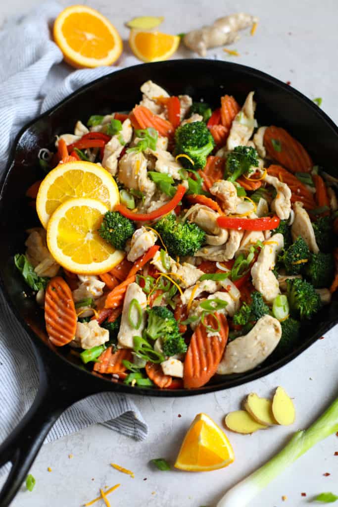 Iron skillet filled with chicken, carrots, broccoli, onions, red peppers and garnished with orange slices