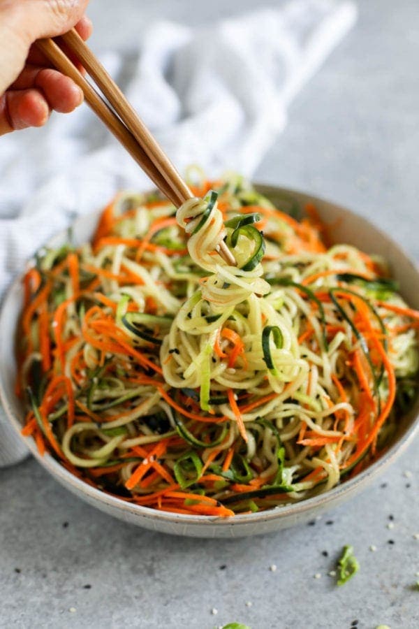 Side view of a white bowl filled with Asian-Inspired Cucumber “Noodle” Salad and a hand using chop sticks to grab a bite