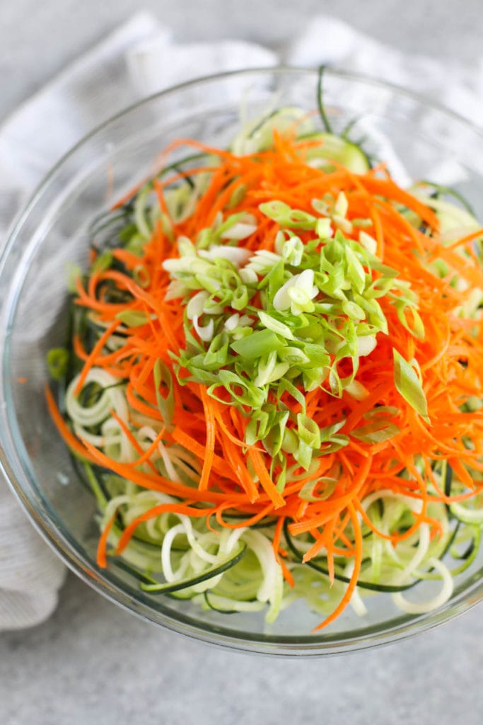 Overhead view of a glass bowl filled with zucchini noodles and carrot shreds topped with green onions