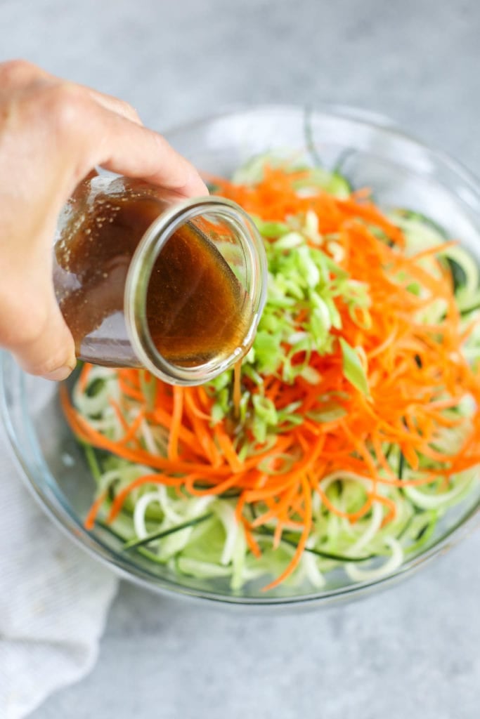 Close up view of a hand pouring dressing over an Asian-Inspired Cucumber “Noodle” Salad in a glass bowl