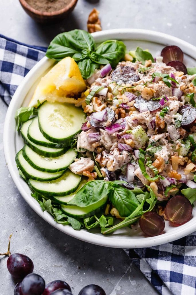 Chicken salad served on a plate of greens with cucumber, nuts, grapes and lemon garnish; checker napkin underneath plate.