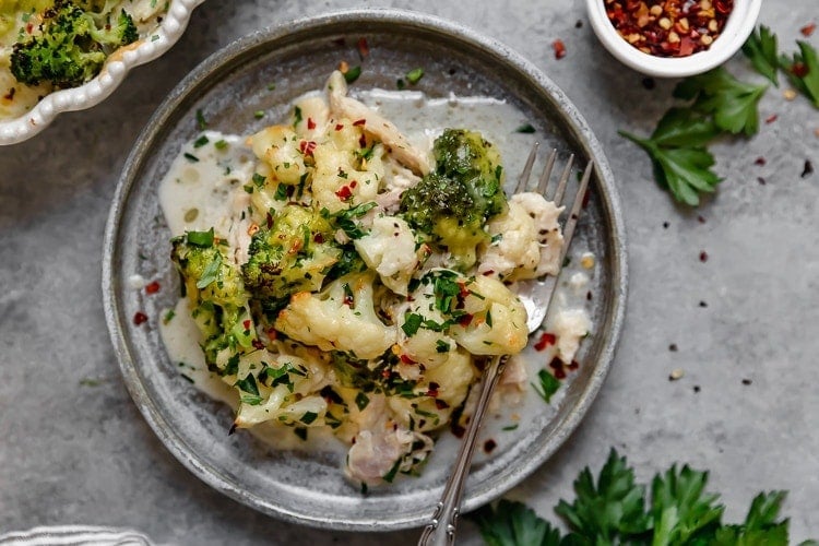 Broccoli cauliflower cheddar chicken casserole serving on grey stone plate, topped with fresh herbs.