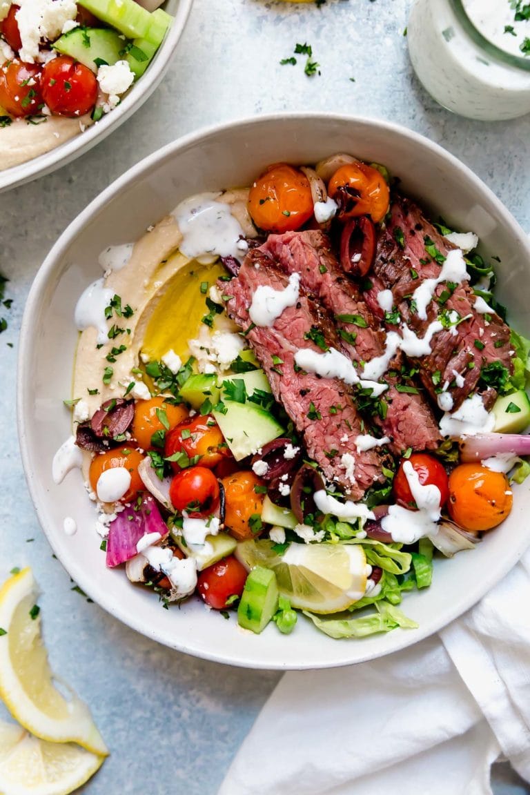 Overhead view of a bowl filled with colorful vegetables, hummus, and sliced grilled steak topped with fresh herbs and a white mediterranean dressing. 
