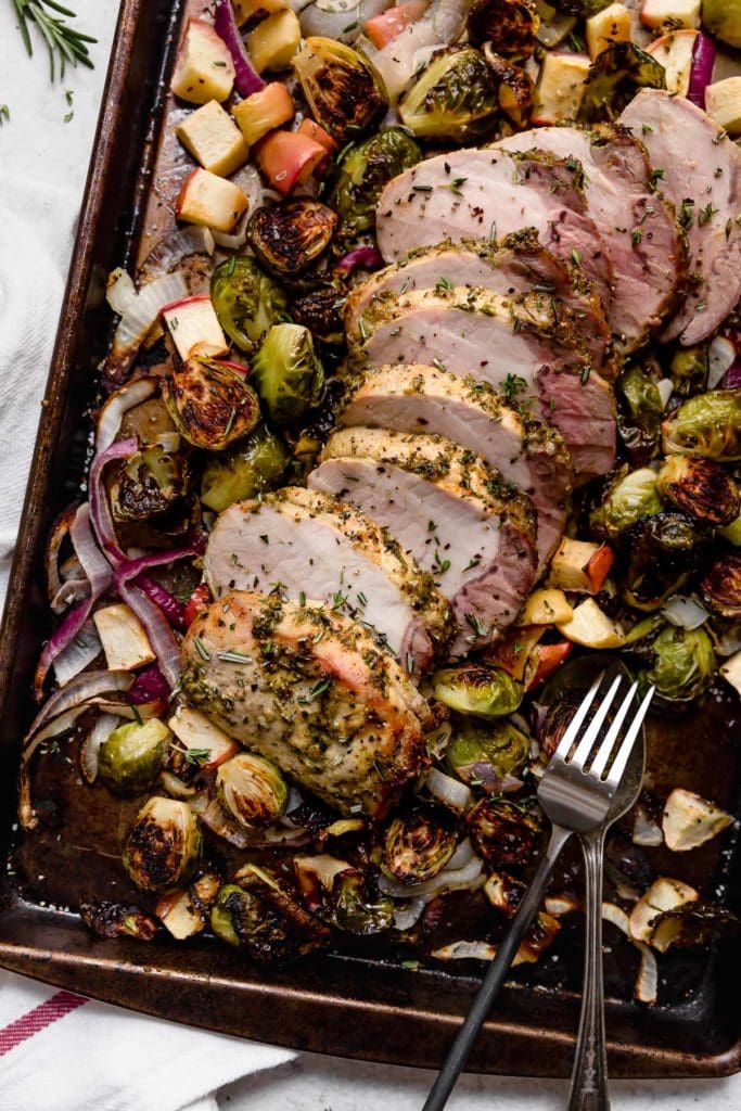 Overhead view of a sheet pan of pork loin with brussels sprouts and apples topped with fresh herbs. 