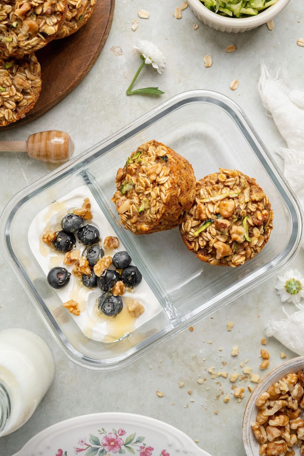 Two oatmeal cups in a meal prep container with a side of yogurt topped with blueberries, walnuts, and a drizzle of honey.