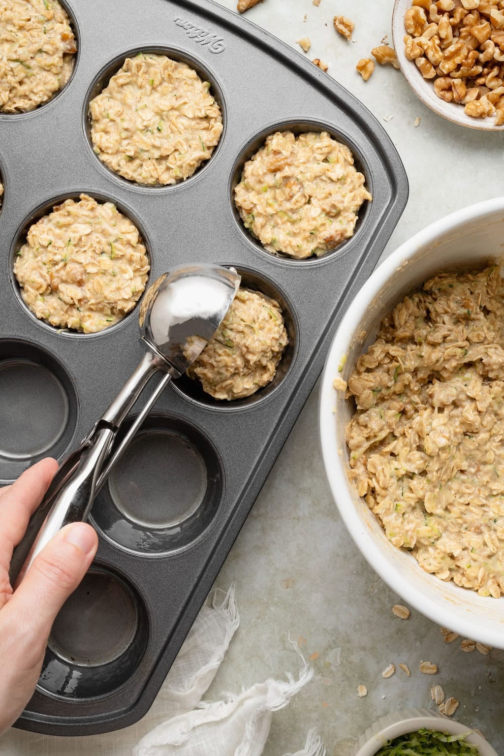 A hand using a cookie scoop to portion oatmeal cup batter into a greased muffin pan.