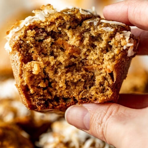 A hand holding up a morning glory muffin with a bite taken from side to show inside texture