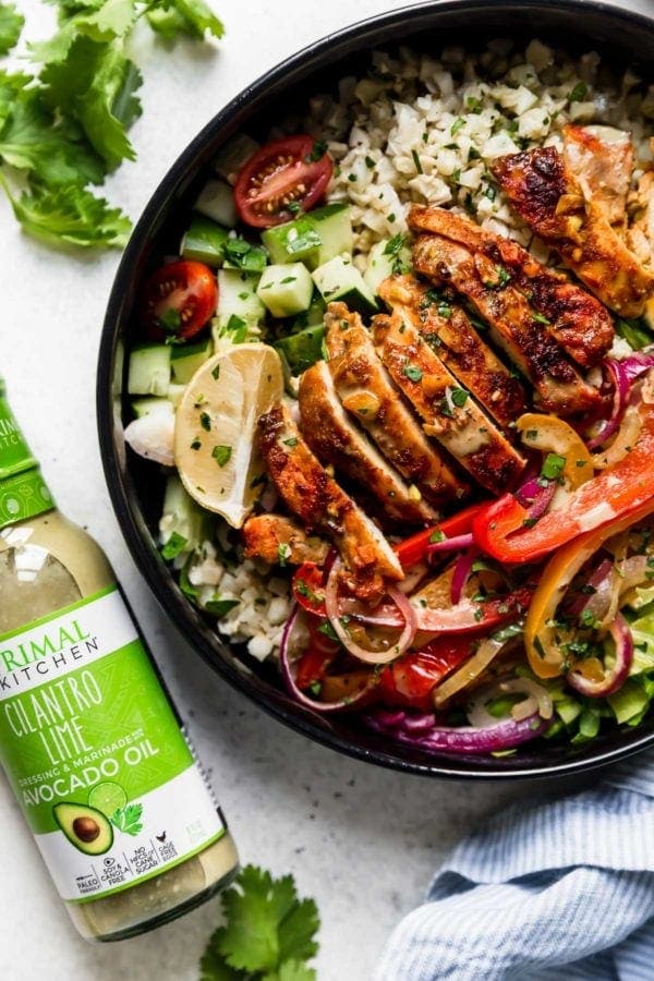 Sheet Pan Chicken Shawarma Bowls: A black bowl filled with cauliflower rice, roasted veggies, sliced chicken thighs, lettuce, cucumbers, and cherry tomatoes and drizzled with a cilantro-lime sauce. 
