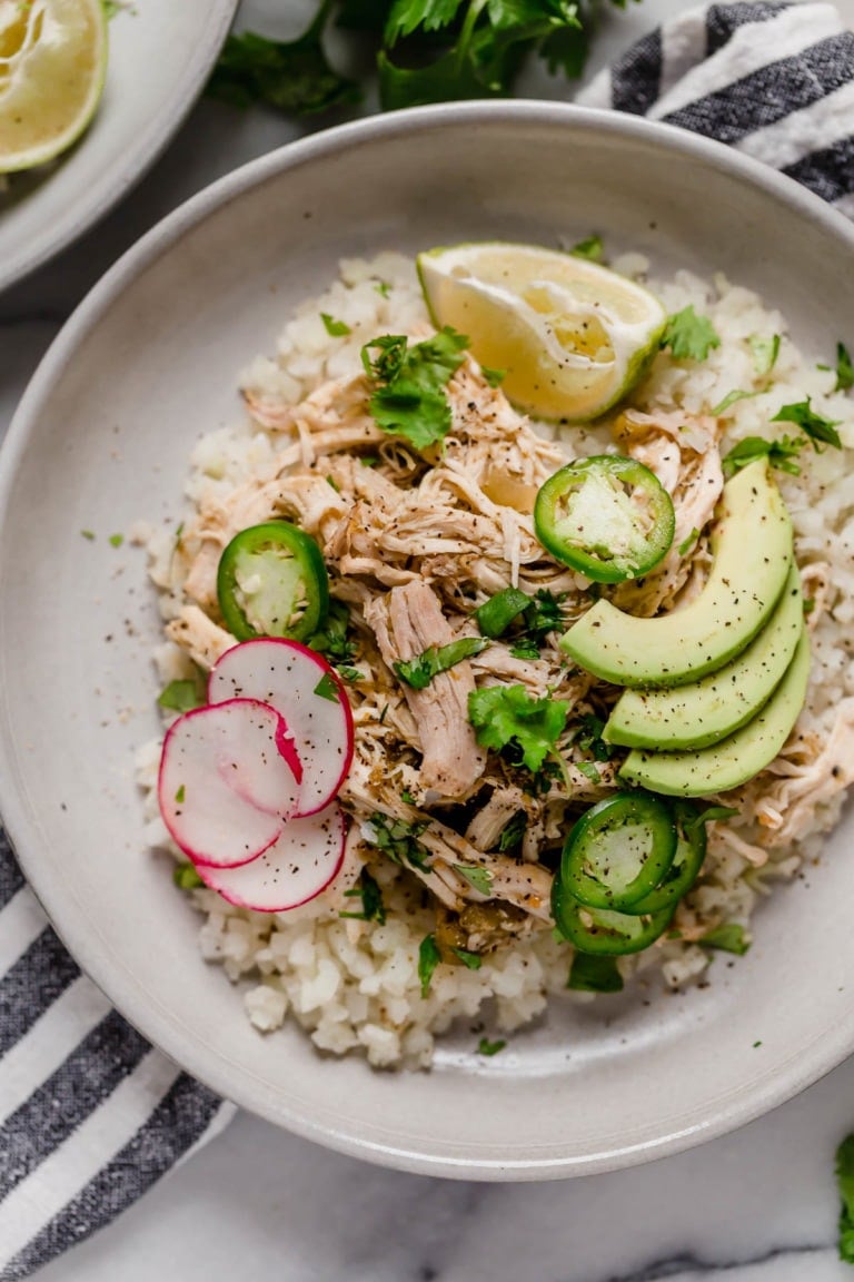 Overhead view of a plate filled with cauliflower rice, saucy shredded chicken, sliced radishes, herbs and avocado.