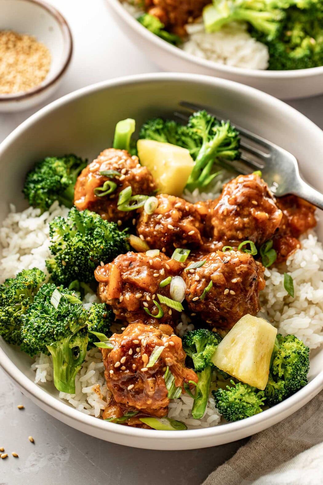 Close up view of a rice bowl topped with sweet and sour meatballs and broccoli garnished with fresh pineapple and sprinkled with sesame seeds and green onions.
