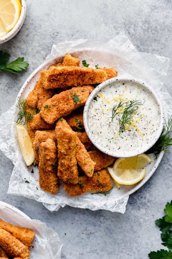 Overhead view of a white plate filled with Baked Fish Sticks with Tartar Sauce topped with fresh dill and garnished with lemon wedges.