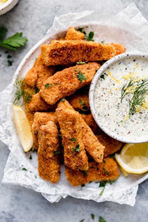 Overhead view of a white plate filled with Baked Fish Sticks with Tartar Sauce topped with fresh dill and lemon wedges.