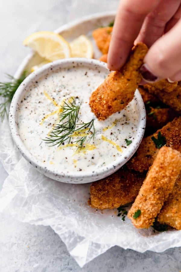 Overhead view of a plate of Baked Fish Sticks with Tartar Sauce and a hand dipping a fish stick in the tartar sauce.