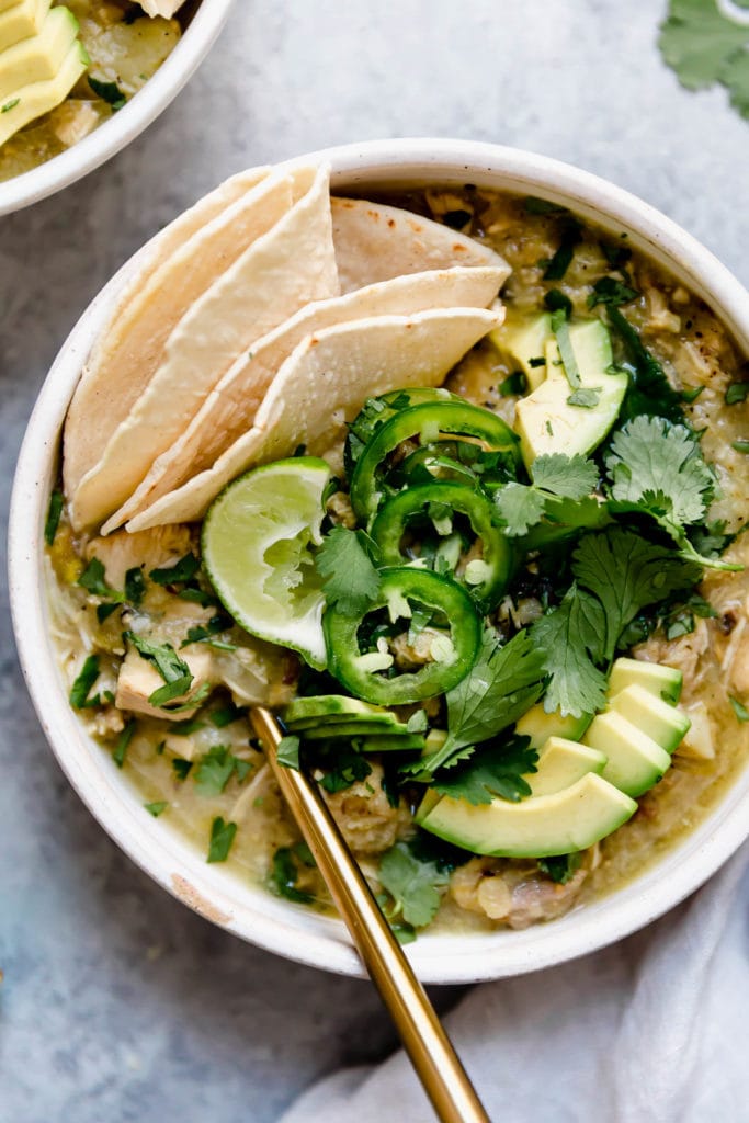 A white bowl filled with Slow Cooker Chicken Chile Verde Stew and topped with fresh cilantro, jalapeño slices, and avocado plus there's a corn tortilla tucked into the side of the bowl for serving. 