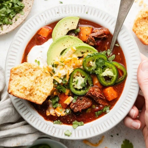 Overhead view of a bowl of Slow Cooker Chuck Roast Chili With Sweet Potatoes topped with a dollop of sour cream and a cornbread muffin on the side.