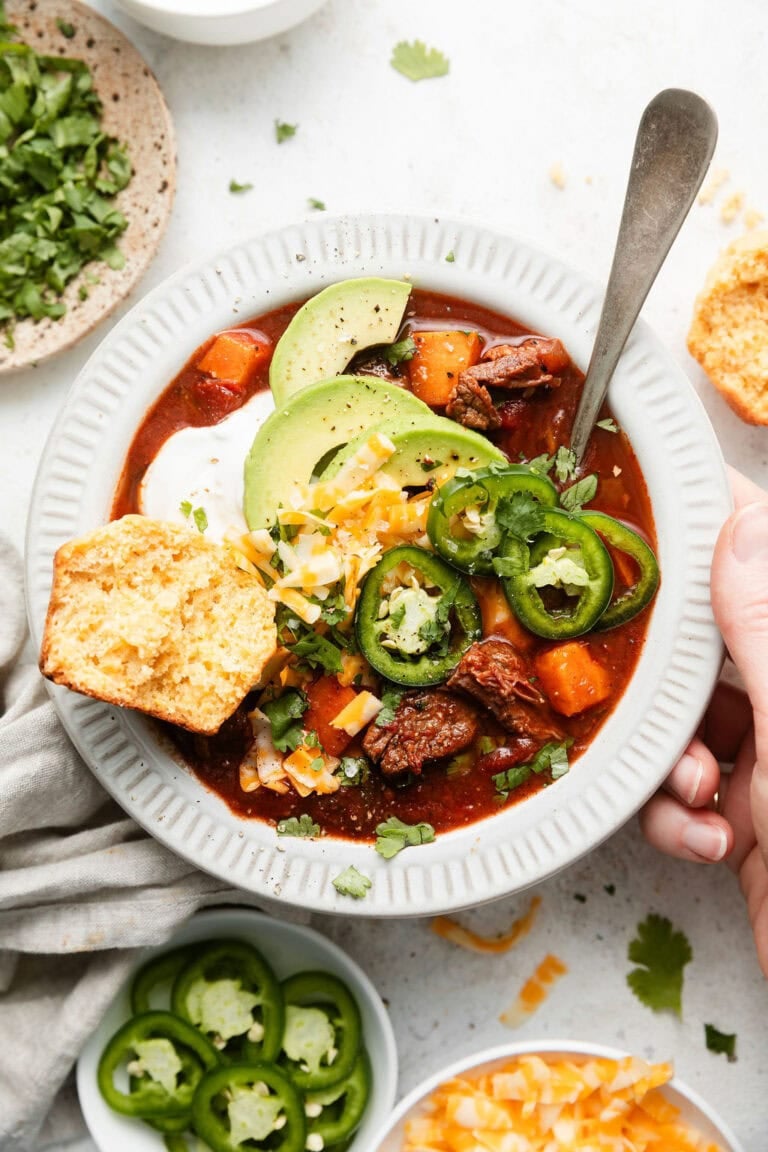 Overhead view of a bowl of Slow Cooker Chuck Roast Chili With Sweet Potatoes topped with a dollop of sour cream and a cornbread muffin on the side.